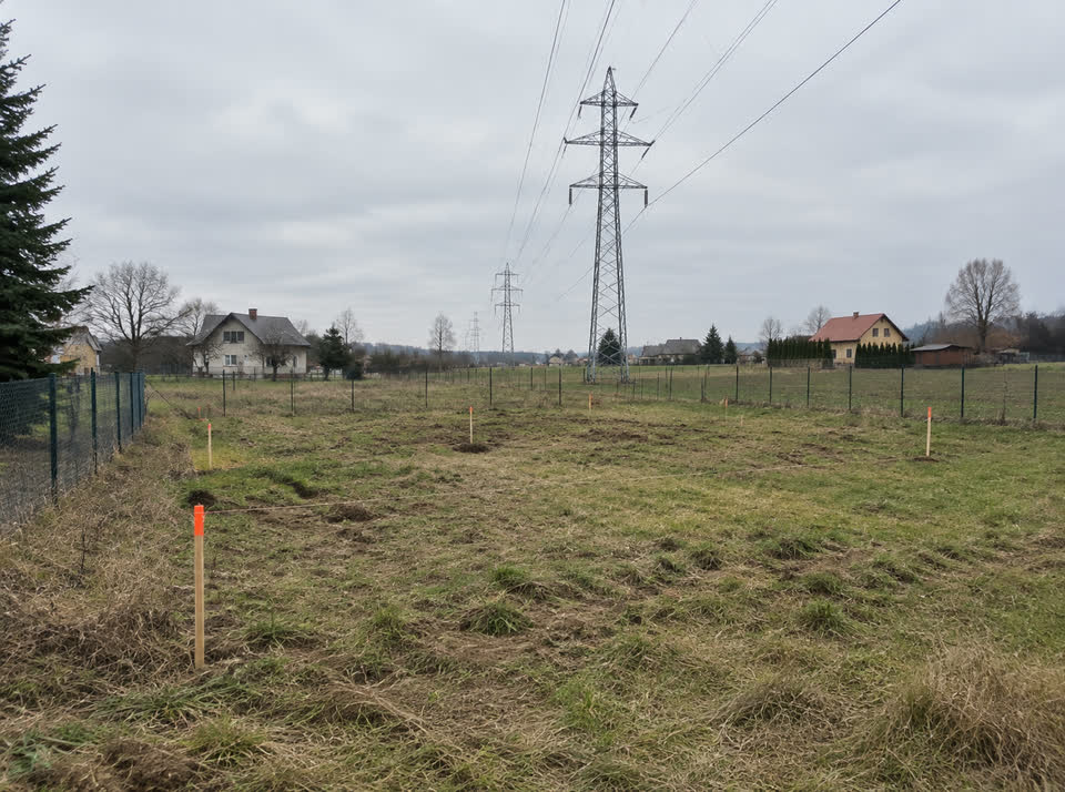 Pusta działka budowlana na wsi ze słupkami geodety i ogrodzeniem z siatki, nad którą przebiegają linie wysokiego napięcia
